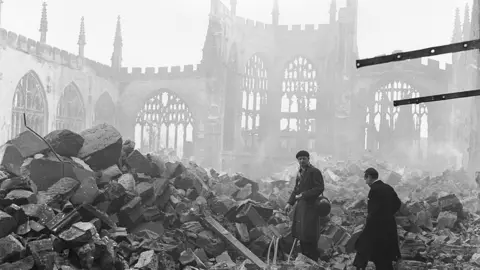 People can be seen amidst the ruins of Coventry Cathedral immediately after the Blitz in 1940. The Cathedral ruins can be in in the background with piles of rubble.