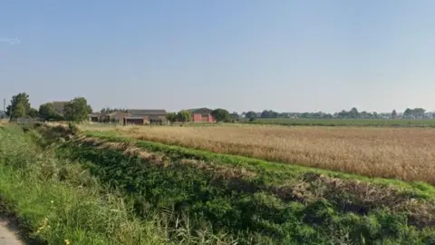 Google A view of the plot from the road. There is a field with a crop growing in it at the moment, with some barns in the background. The weather is sunny.