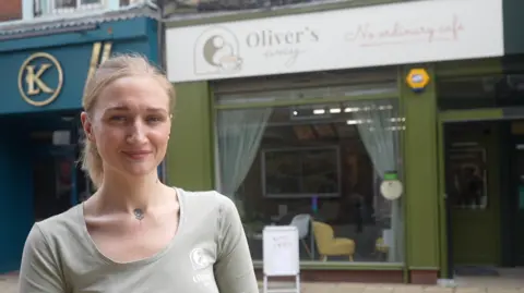 Jamie Niblock/BBC A woman with blonde hair that has been tied back smiles at the camera. She is standing in front of a cafe with a green front. She wears a light green long sleeved top. 