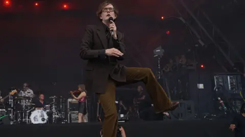 A man in chord trousers and a brown suit jacket is on stage holding a microphone. He has his left leg lifted up. Behind him are musicians on drums and violin.