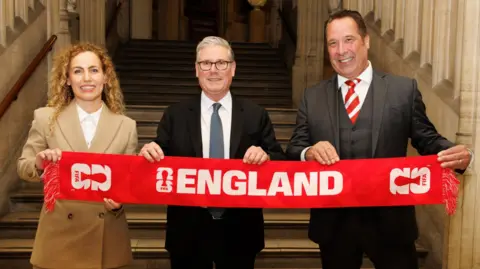 No 10 Downing Street Katie White, MP for Leeds North West, Prime minister Sir Keir Starmer and former England goalkeeper David Seaman stand in a line holding up a red and white England scarf.