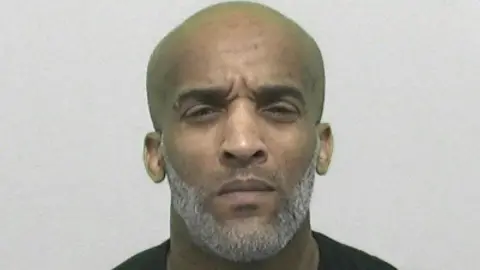 Mugshot of Mark Branker. His head is large and shaved and he has a neat grey beard. He is squinting slightly.