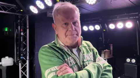 Steve Jason is standing on a small stage which has stage lighting on its ceiling and walls. Jason has short white hair and he is wearing a white T-shirt, and a green jacket which has white writing and stripes.