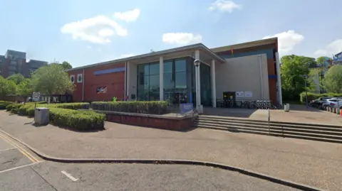 The front of Riverside Leisure Centre. It is a red brick building with glass panels and columns by the entrance. Steps and a ramp out outside the building.