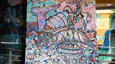 Trivandi An abstract, expressionist painting of bright colours that show Glasgow's landmarks. There is a bridge, a crane and a statue painted amongst other abstract pictures. The painting is on an easel central to the photograph.