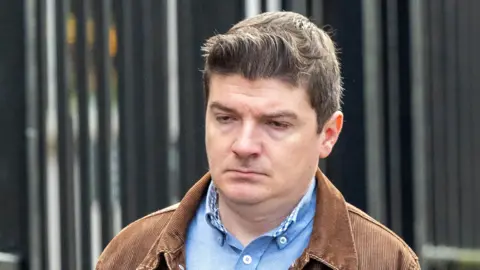 Pacemaker Michael McMonagle walking into court  wearing a blue shirt and brown jacket. His hair is short and brown. The courthouse railings can be seen in the background 