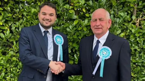 Robin Hunter-Clarke is shaking hands with Julian Kirk. The pair are both wearing navy suits, blue ties and Reform UK rosettes. Hunter-Clarke has a blue and white pin-stripe shirt, whilst Kirk has a white shirt on.