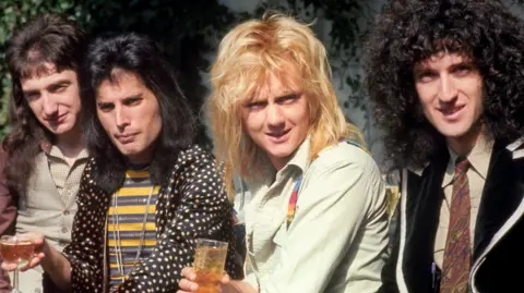 PA Media Queen from left: John Deacon has long brown hair, he is wearing a light brown shirt with checked black lines and a similar waistcoat; Freddie Mercury is holding a glass of champagne in his right hand, has long black hair, a black jacket with yellow spots and T-shirt with alternating black and yellow horizontal stripes; Roger Taylor has long blonde hair and is wearing a light green shirt with rainbow-coloured braces; Brian May has long black curly hair and is wearing a black blazer with white edging like Patrick McGoohan in The Prisoner, a light khaki shirt and brown Paisley patterned tie.