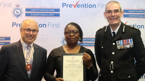 Nancy Segwete holding her award from Judge Michael Kay and Chief Constable Charlie Hall at Hertfordshire Constabulary police headquarters