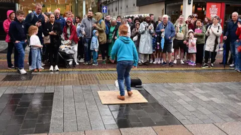 Pacemaker A young ginger boy dancing on the pavement for a large crowd