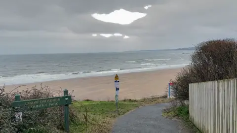 Photo of Helen's Bay beach. The tide is out and sand can be seen. The sky is grey and the water looks calm.
A beach is visible near a sign reading: Crawfordsburn Country Park