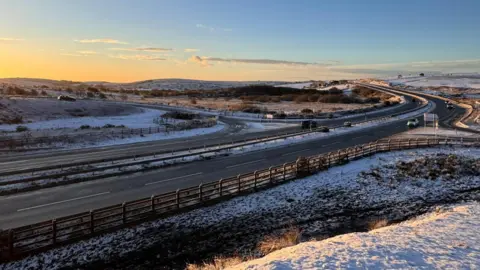 The A30 at Temple, Bodmin is surrounded by snow at sunrise. The lanes have just a few vehicles.