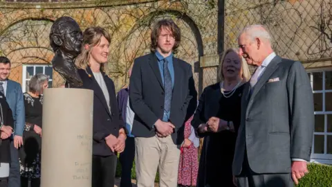 The King's Foundation King Charles views a bust of his head alongside the sculptor - a woman with tied back blonde hair who is smiling - and two other people, a man with long hair and glasses wearing a suit jacket and chinos and a woman with blonde hair wearing a black dress and pearls. 