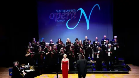 John Lloyd Davies A full opera performance on a theatre stage featuring a large choir arranged on tiered risers in the background. A soloist in a red dress is standing at the front. A conductor on a small podium is facing the performers and a pianist is seated at a grand piano to the side.