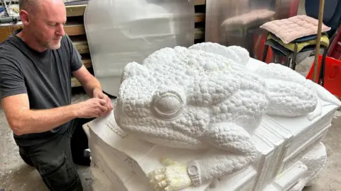 Simon Thake A man kneels down next to a large polystyrene model toad.