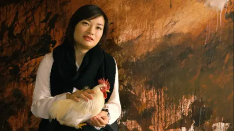 Q Films A person sits in front of a large, abstract backdrop with earthy brown and black tones.
The person holds a light‑coloured chicken in their lap, with one hand resting gently on the bird.