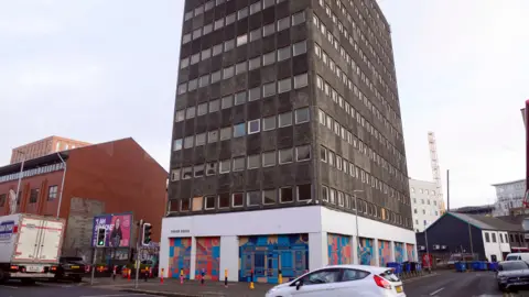 Pacemaker Grey tower block with some of it's windows boarded up. On the ground floor, which is also boarded up, there is some colourful artwork of buildings. Outside the buildings is a road with some cars, a lorry, and traffic lights. Next to the building is a billboard.