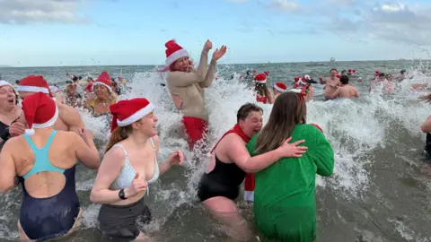 A large ocean wave hits a crowd of woman wearing swim suits and red Santa hats with white tips. There are many more people wearing swimsuits beyond the wave.