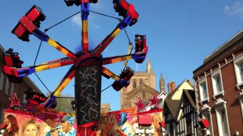 A large fair ride with spinning carts is pictured on a street with old buildings on either side of it and a large cathedral in the backdrop.