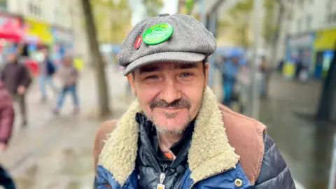 DAVE EDWARDS/BBC A 43-year-old man looks into the camera. There's a badge on his hat which reads "Act now on day services for rough sleepers in Doncaster".