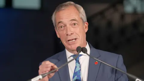 PA Media Nigel Farage giving a speech at Banking Hall in the City of London. He is wearing a blue suit and tie with a red poppy pin badge and speaking into a microphone.