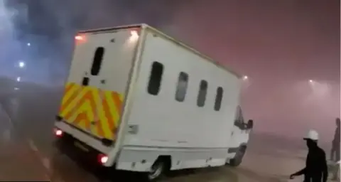 A screenshot of the police van seen on the Elbit Systems UK site. The van is a large white vehicle with small windows and a chevron pattern across the back. A figure wearing black with a white helmet can be seen in the lower right corner of the image holding a long unidentified object.