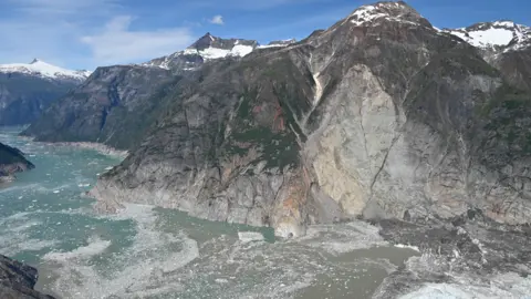An icy expanse of sea runs through a narrow passage between mountains. Much of the steep sides of the mountains are stripped of green vegetation revealing grey scarred rock. The tops of the mountains are scattered with snow and ice glitters in the fjord below. 