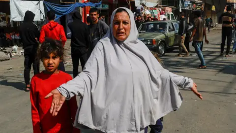 A woman in a grey hijab stands with a child holding her hands out in dismay in Rafah in the southern Gaza Strip March 18, 2025