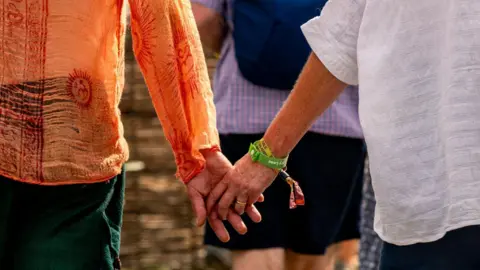 Garry Jones Photography Two festival-goers, one wearing an orange sun-print shirt and the other wearing a white shirt, hold hands as they walk through the site. The person on the right has two festival wristbands on, one yellow with a pink pattern and another overlapped over the top in a solid green. 