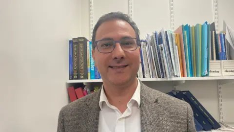 Ellen Knight/BBC Prof Rakieh is pictured looking into the camera and smiling. He's wearing a white collared shirt, and a light brown tweed jacket. He's also wearing a pair of dark blue-framed glasses. He's stood inside an office with white walls. Behind him are two shelves of books, files, and folders. Over Prof Rakieh's right shoulder are two dark brown books with the word 'RHEUMATOLOGY' written clearly on them. 
