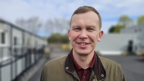 Michael Bradley is smiling at the camera. He has light brown hair and is wearing a red shirt underneath a khaki green coat.