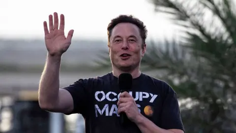 Elon Musk waving towards the camera wearing a T shirt with the slogan Occupy Mars