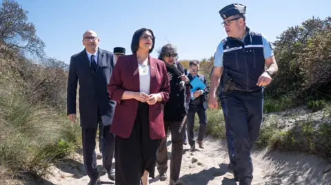 PA Media Home Secretary Shabana Mahmood is wearing a black skirt, a white top and a maroon jacket. She is walking along a sandy path next to a French law enforcement officer. There are four people walking behind her including French Interior Minister, Laurent Nunez who is wearing a suit. 