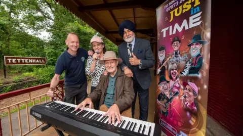 Three me and four women stand on a balcony surrounded by railings, with a sign saying Tettenhall in the background. A man in the centre in a hat, brown leather jacket and green shirt is sat with his fingers on a keyboard. A women behind his right shoulder in a black-and-white spotted shirt holds a microphone to her mouth. A man on her right is wearing a dark polo-shirt and trousers and posing with his thumb up; on her left is a man in a suit with a grey beard, wearing a black turban.