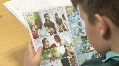 A boy reads from a comic he is holding above a table. He is pictured from behind, and some of the action on the page is visible, and involves conversation between two male characters.