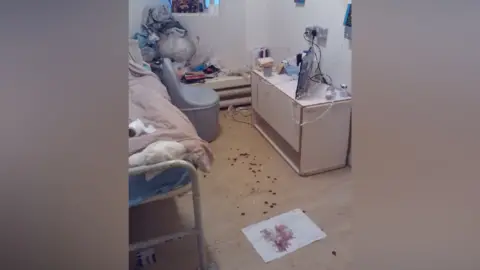 HM Inspectorate of Prisons A small, cluttered prison cell with a bed, chair, and desk which is strewn with rubbish and personal items, while dark blood stains are scattered across the floor and a sheet of paper, suggesting a recent disturbance.