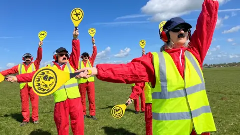 A group of people are dressed in red jumpsuits with yellow high-vis vests and they are - in unison - holding up yellow air traffic control signals which show images of the world burning. They are standing in a field.