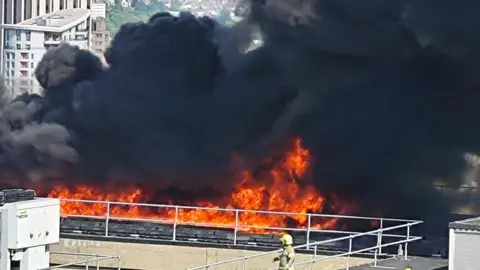 Marina Ribeiro Kaufmanner Firefighter stood in front of flames on the roof of the building. Black plumes of smoke are rising behind the orange flames.