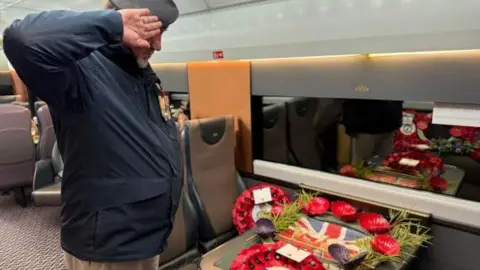 Cornwall Partnership NHS Foundation Trust Jason Thomas saluting and placing a Trust wreath on the Poppies to Paddington train at Penzance station. The wreath is on the train table. It is the inside of the night riviera sleeper train. 