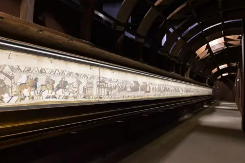 Bayeux Museum Photo shows A long section of the Bayeux Tapestry  which is shown in a darkened space in which only the artefact itself is lit.
