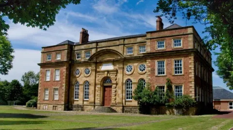 Kirkleatham Museum is a grand three-storey brick and stone building in a grassy field. There is an arched window above the tall brown door. The other windows are a mix of circular and square designs. A lush green vine grows on the front wall.
