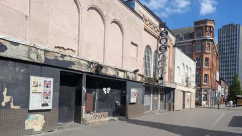 A building in a poor state of repair with a wide pavement in front and blue sky above