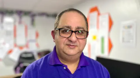 BBC/Martin Giles A man in wearing glasses and a purple care home shirt sits at an office desk.