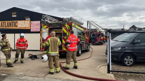 Owen Sennitt/BBC There is a fire engine with hoses trailing from it and another fire engine with an aerial platform behind it. Fire fighters are standing around the fire engine in a yard. There is a commercial building with the sign: Atlantic Truck and Vehicle Centre.