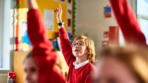 Getty Images a 10 or 11 year old boy raises his hand in a classroom full of other students with their hands up. They are all wearing red school uniforms and the boy - who is the only one in the picture - is 