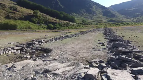 Lots of rocks are visible and a small pool of water. The rocks run parallel and there are hills and greenery in the background.