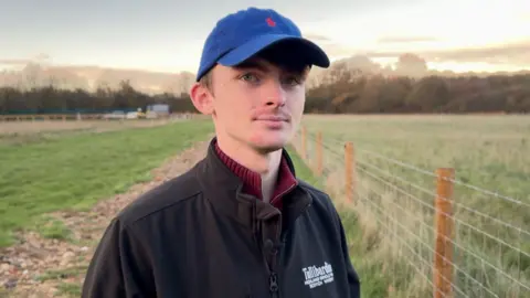 Joshua wears a blue cap and a black fleece. He stands by the fence at the edge of his field.