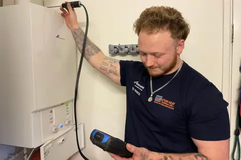 A man with ginger hair and a tight blue T-shirt checks out a gas boiler with a probe 