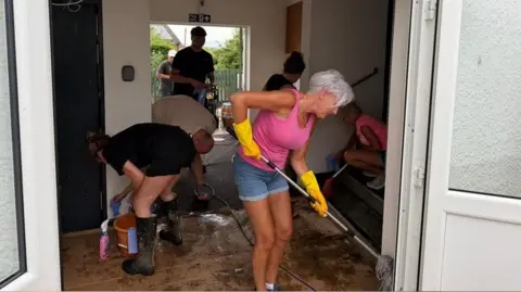 Several people use mops, cloths and buckets to clean floors and walls in the lobby of a building. The floor appears to have black char marks.