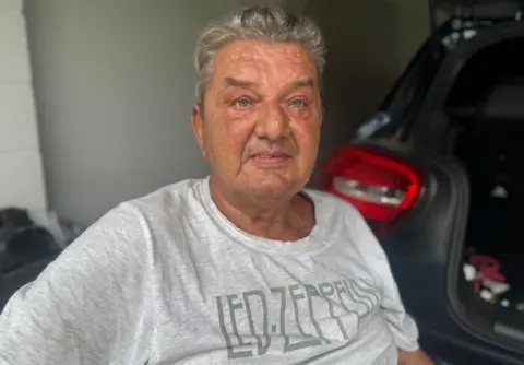 An Older White Man, Wearing A Gray T-Shirt with Led Zeppelin Written Across the Front, Sits in Front of a Car In What Appears to Be a Private Garage